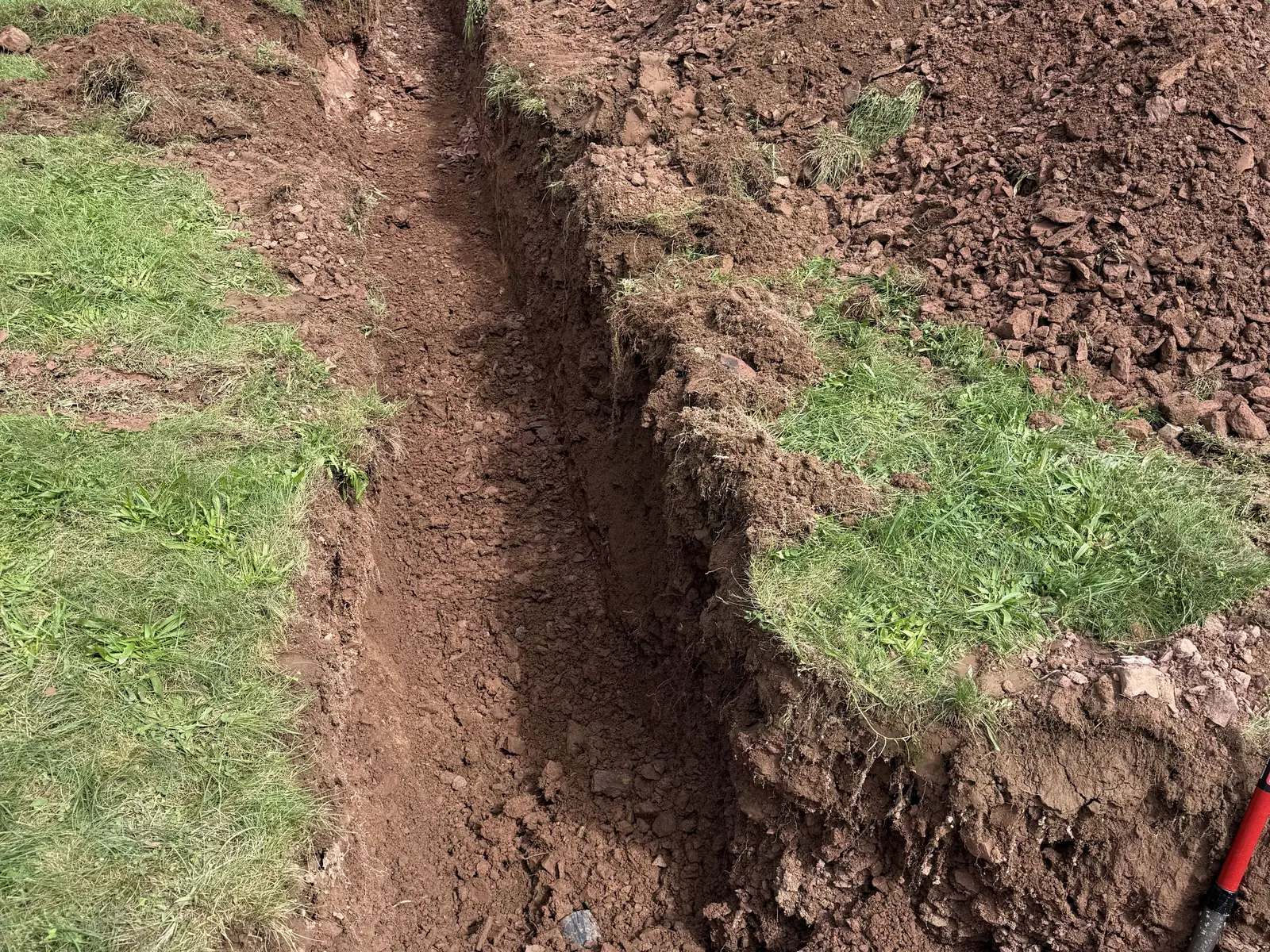 Narrow trench cut through rocky red soil, clean walls and the spoil beside it
